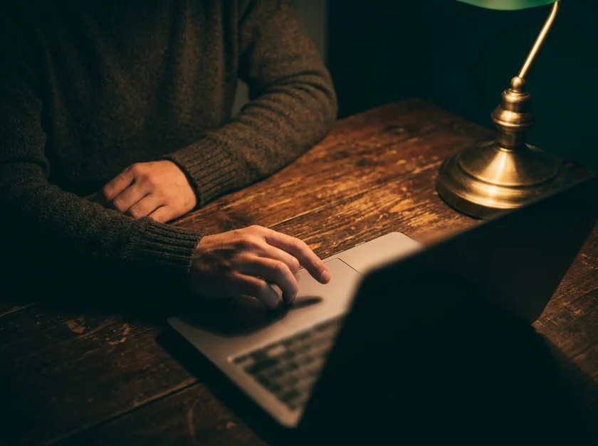 Person in a dark sweater reviewing work on a laptop at a wooden desk, lit by the warm glow of a green banker's lamp